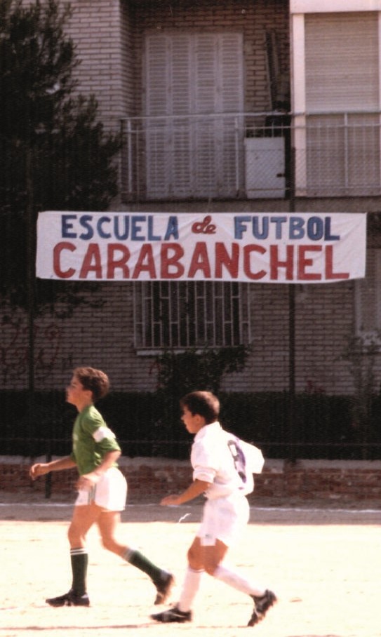 dos niños jugando al fútbol con una pancarta de fondo que se lee Escuela de Fútbol de Carabanchel