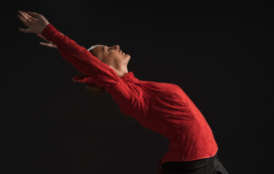 Una bailaora de flamenco con un traje rojo sobre fondo negro estirando los brazos hacia atrás