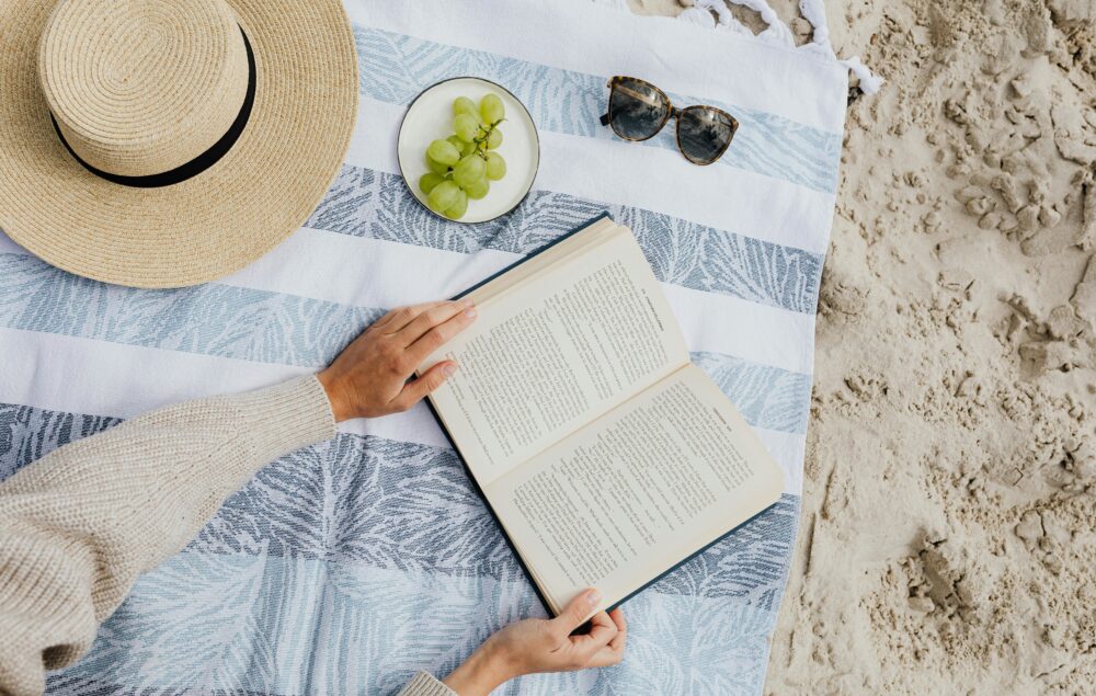 fotografía de una mujer leyendo un libro sobre una toalla, con un sombrero unas uvas y unas gafas de sol