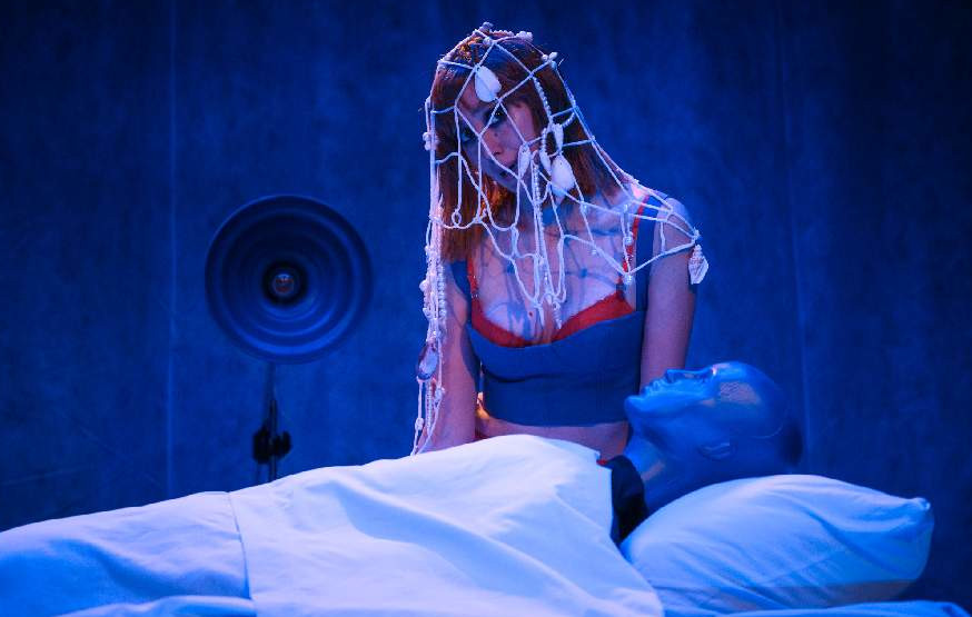 Fotografía de la obra Suavecita. Una mujer mirando a un hombre en una cama de hospital con tonos azules