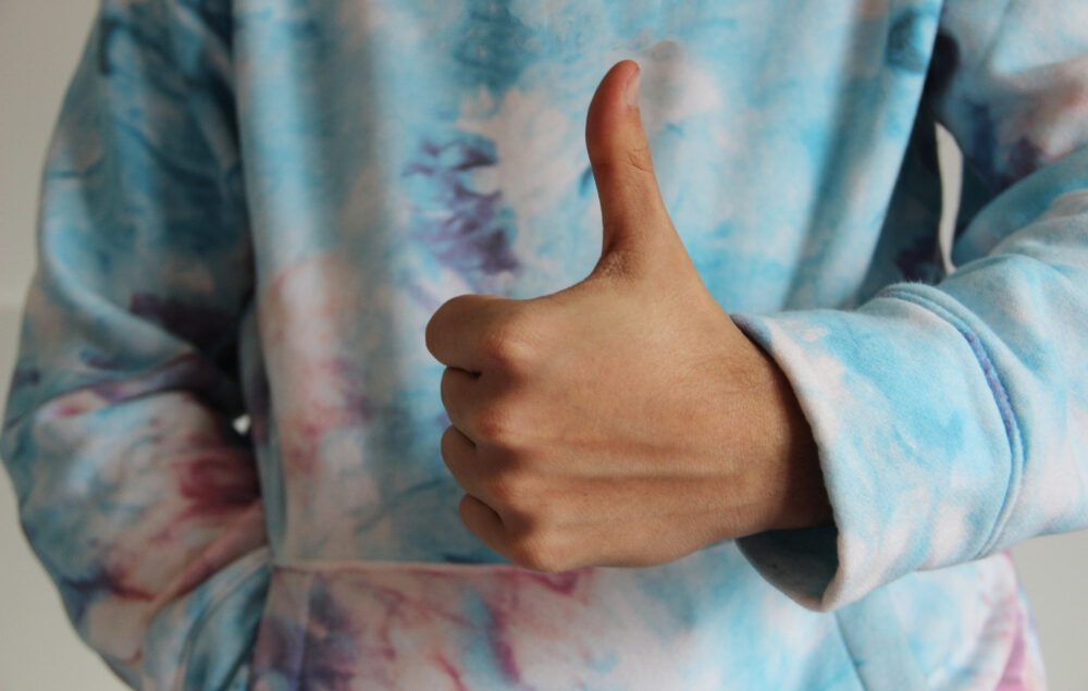 Una mano con el pulgar hacia arriba en señal de aceptación. Se ve una sudadera colorida por detrás