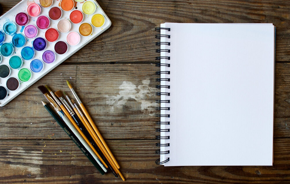 acuarelas y pinceles junto a un cuaderno en una mesa de madera
