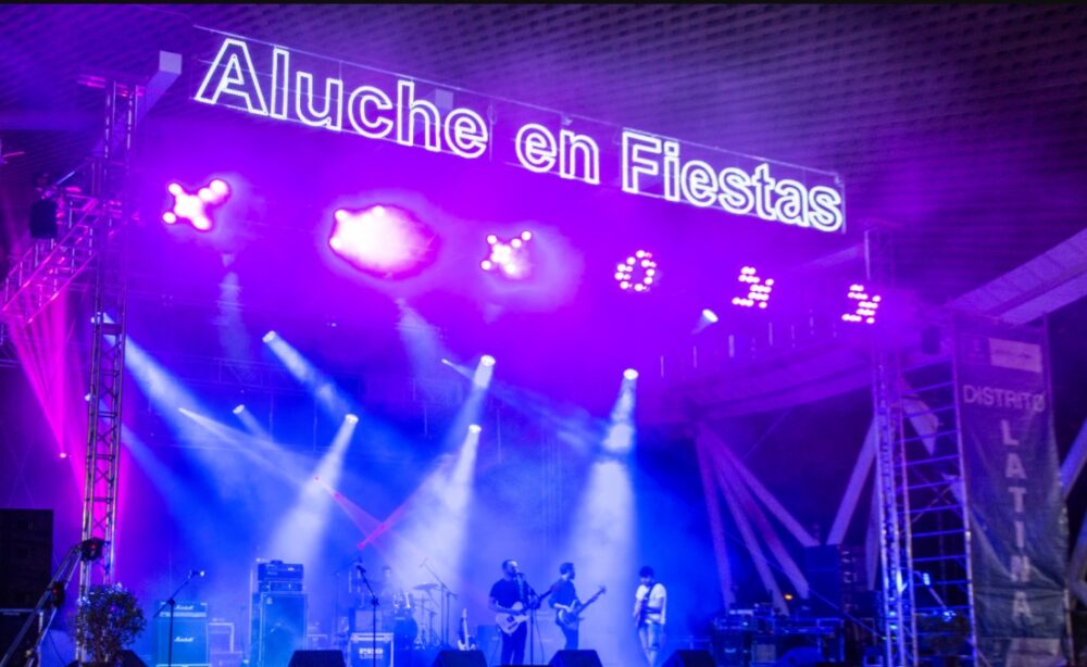Escenario nocturno de las fiestas con una actuación musical y un lema luminoso de Aluche en Fiestas