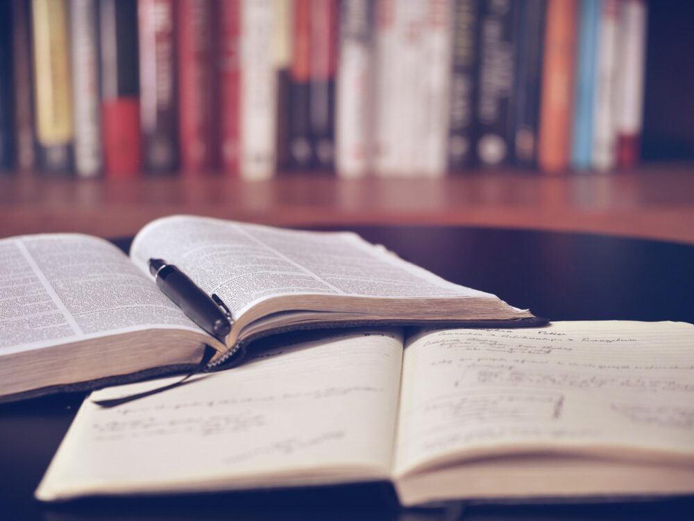 Libro con boli y cuaderno abiertos en mesa de biblioteca con estantería con libros de fondo