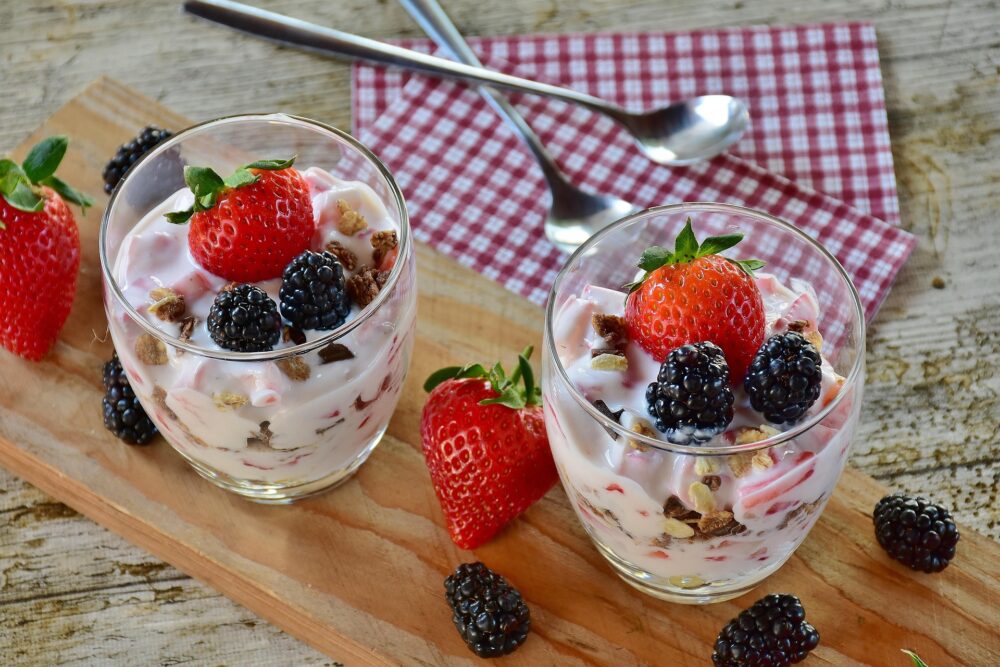 Foto a dos tarrinas de cristal con yogures con fruta y cereales sobre una mesa de madera con servilletas a cuadros vichy rojos y blancos y dos cucharillas largas