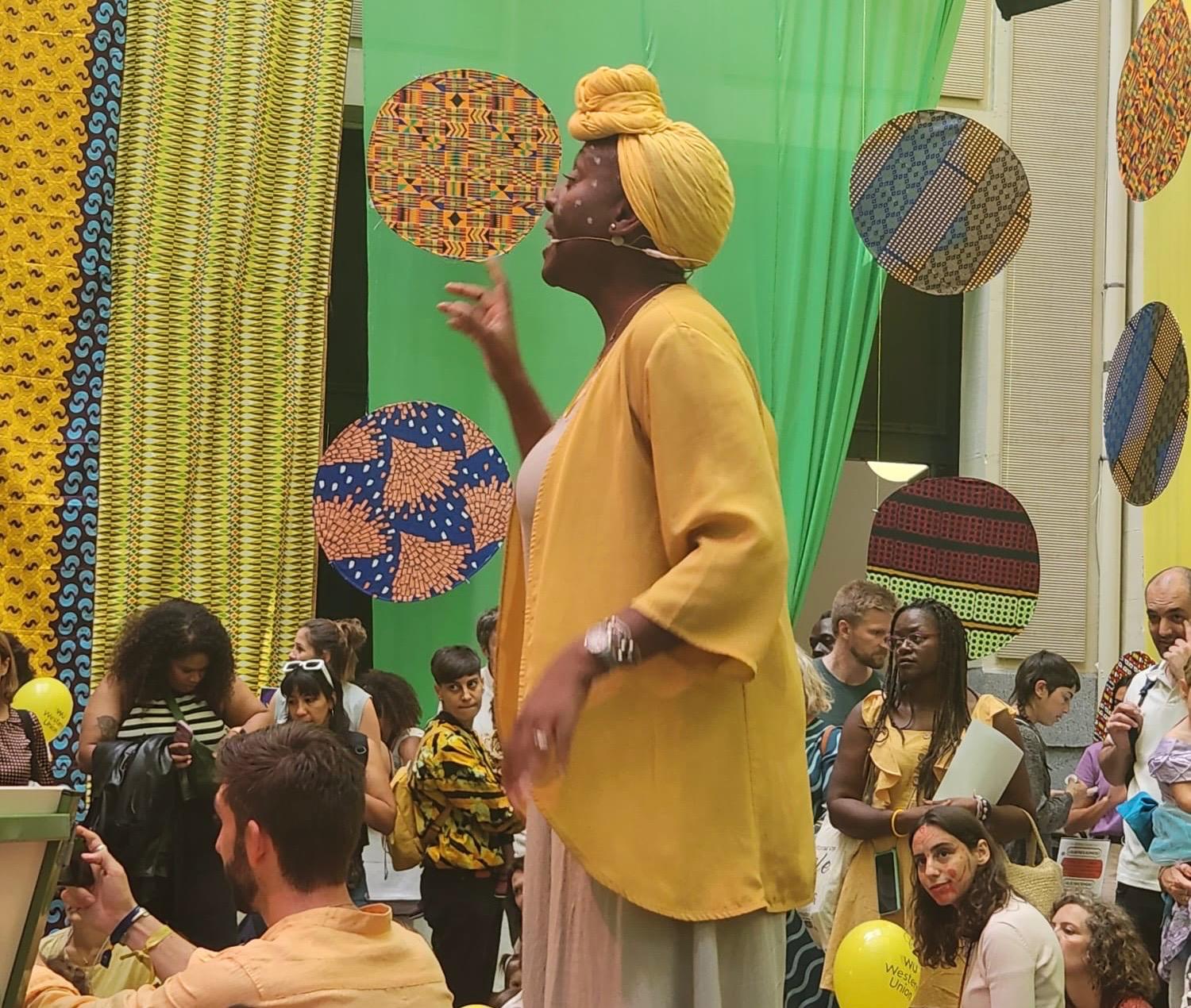 Foto promocional deBokobo Kids Village donde aparece de pie una cuentacuentos africana vestida de amarillo con público