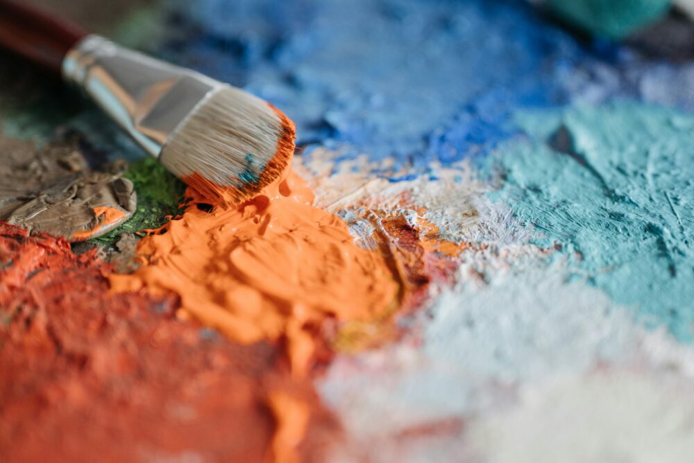 Brocha cargada de pintura naranja sobre una paleta con mezclas de colores vibrantes como azul, turquesa, rojo y marrón, en una escena artística de primer plano