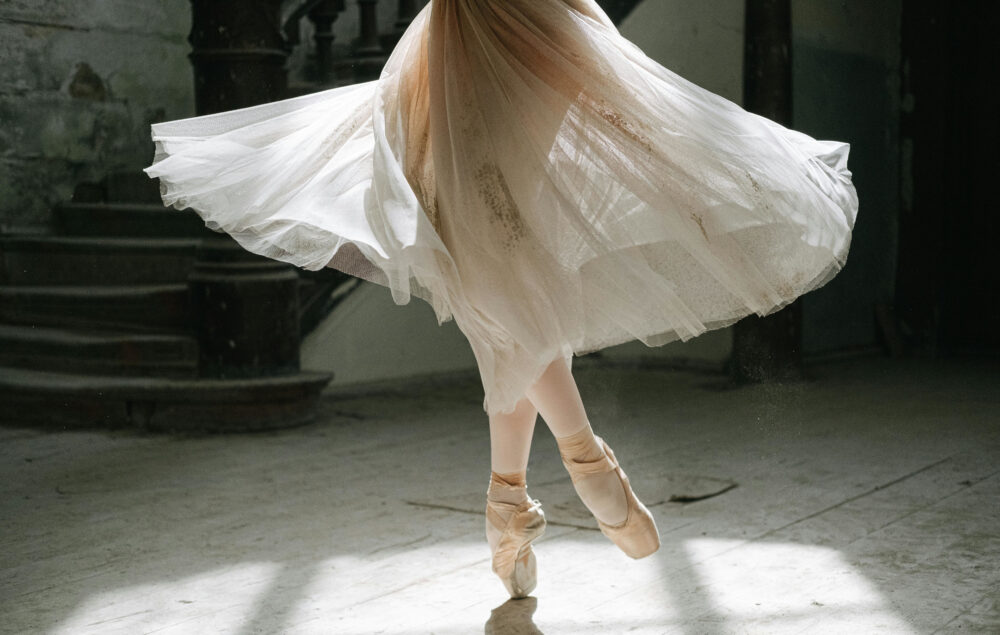 Bailarina de ballet en puntas girando con un vestido vaporoso blanco, captada en pleno movimiento en un espacio antiguo con suelo de madera y escaleras al fondo.
