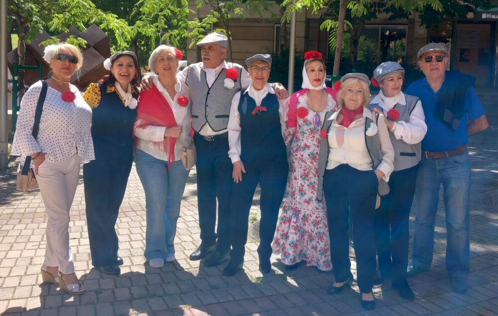 Grupo de personas mayores posando al aire libre durante la celebración de San Isidro, varias de ellas vestidas de chulapos y chulapas con claveles rojos y mantones, en un ambiente festivo y soleado.