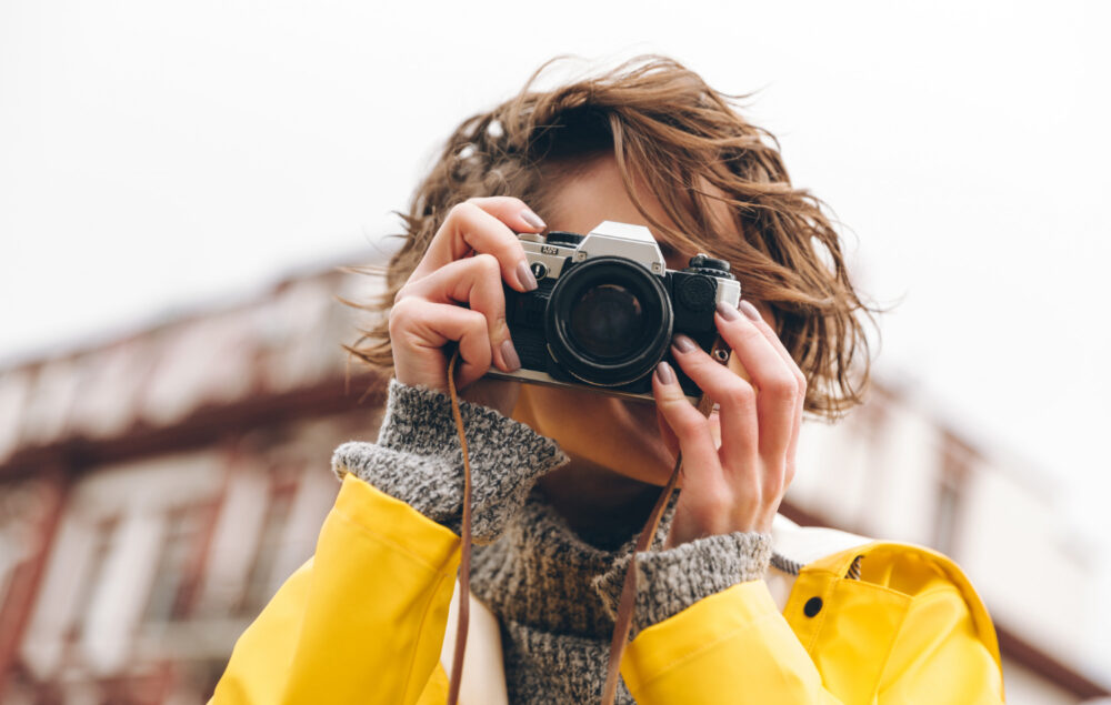 Una mujer realizando una fotografía con una cámara analógica. lleva un chubasquero amarillo y el pelo corto en un día claro
