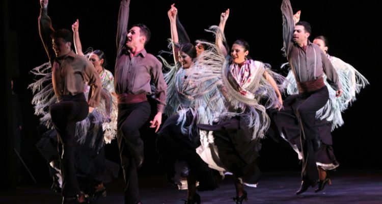 Grupo de hombres y mujeres bailando flamenco sobre un escenario, fondo negro