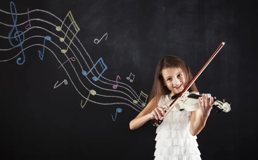 Niña sonriente tocando el violín frente a una pizarra negra con notas musicales de colores dibujadas con tiza.