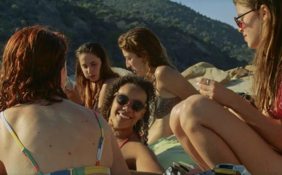 Grupo de jóvenes en traje de baño sentadas en círculo sobre una toalla en la playa, conversando y disfrutando bajo el sol, con colinas boscosas al fondo.