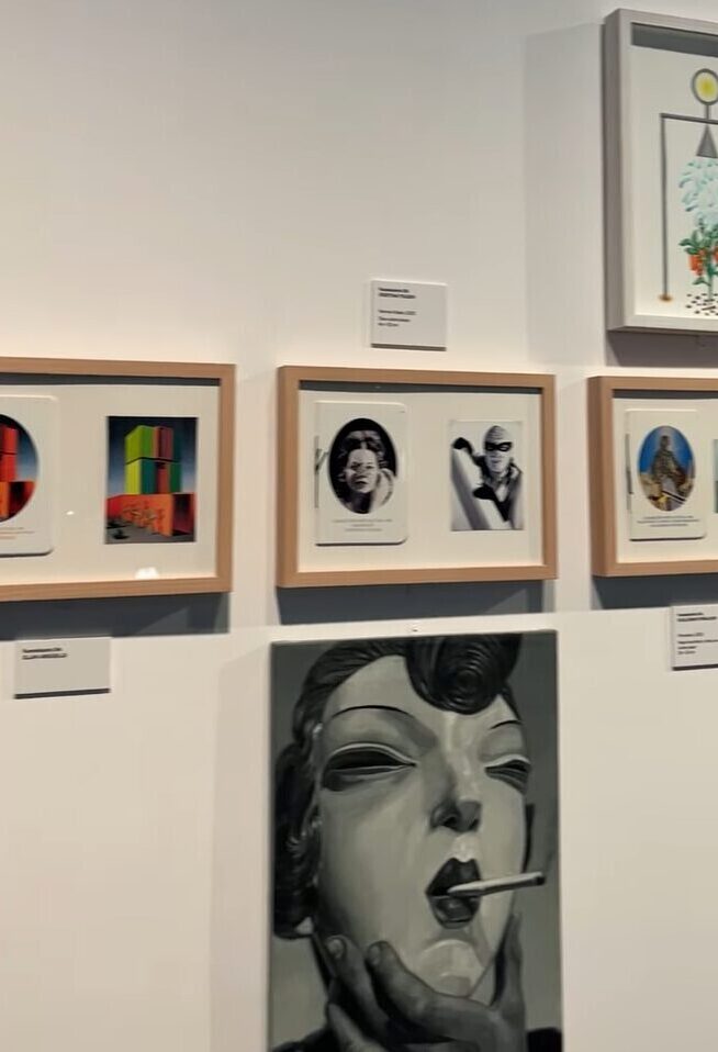 Vista parcial de una pared de galería con varias obras de arte enmarcadas, incluyendo collages, retratos y una pintura en blanco y negro de un rostro femenino con un cigarrillo en la boca, sujeta por una mano.