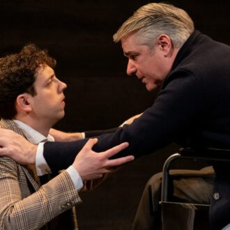 Escena teatral con dos hombres mirándose intensamente; uno de ellos está de rodillas frente al otro, que se encuentra en una silla de ruedas, mientras ambos se toman de los brazos en un gesto cargado de emoción.