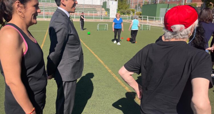 El concejal de Villaverde, Orlando Chacón, durante las olimpiadas, con traje en un campo de fútbol con mayores a los lados con ropa deportiva
