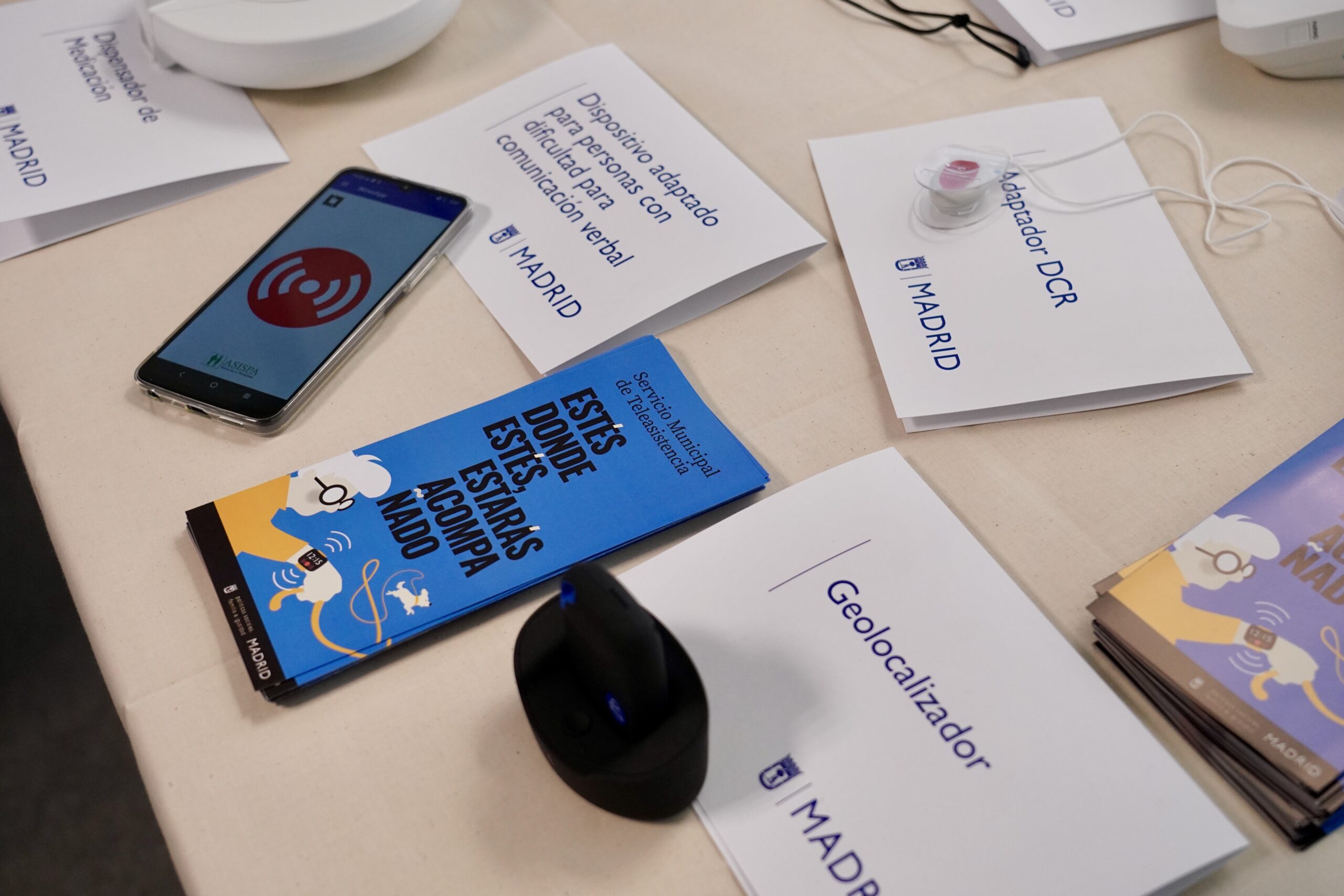 Mesa con varios dispositivos del servicio de teleasistencia del Ayuntamiento de Madrid, incluyendo un móvil con la app de emergencia abierta, folletos informativos, un geolocalizador y un adaptador DCR