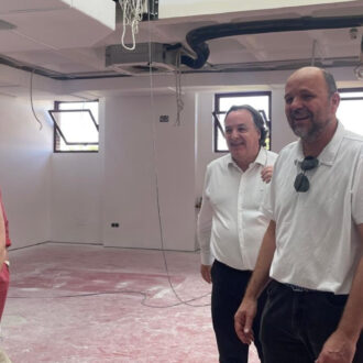 Grupo de personas vestidas de manera formal conversando y sonriendo en el interior de un edificio en obras con suelo cubierto de polvo y techos aún sin terminar.