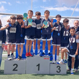 Grupo de niños atletas sobre un podio de premiación al aire libre, con medallas y trofeos, acompañados por varios adultos; fondo con red de portería y banderolas del distrito de Vicálvaro