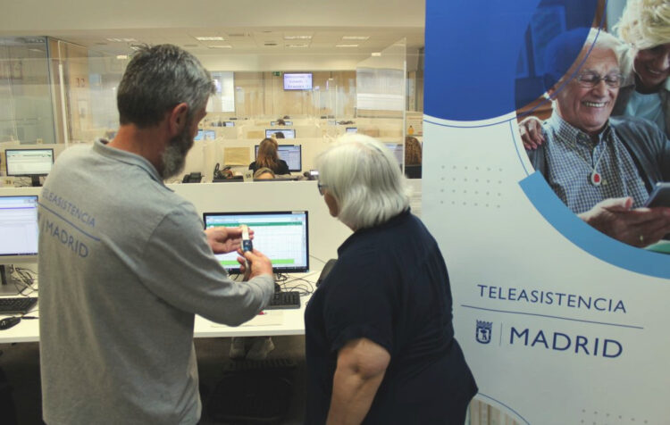 Trabajador del servicio de teleasistencia del Ayuntamiento de Madrid muestra un dispositivo a una mujer mayor en una oficina con operadores al fondo y un cartel promocional del servicio