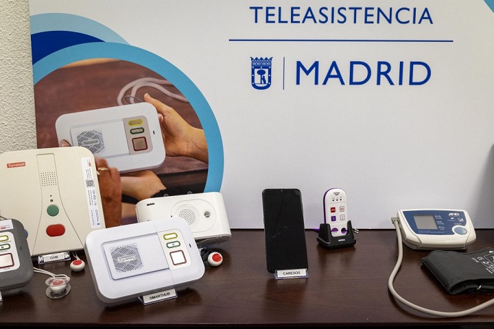 Mesa con varios dispositivos de teleasistencia del Ayuntamiento de Madrid, incluyendo pulsadores, centralitas, teléfonos y un tensiómetro, frente a un cartel institucional