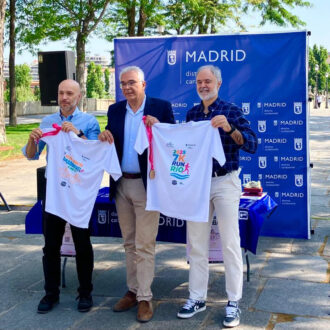 El concejal de Carabanchel presentando la V Media Maratón sujetando las camisetas blancas de la prueba con un cartel azul detrás cerca del Puente de Toledo