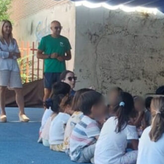 niños sentados en el suelo junto unos adultos de pie, en el campamento de Ciudad Lineal
