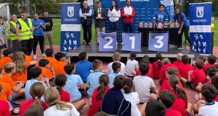 Participantes como público de la entrega de trofeos de las Olimpiadas Escolares de Fuencarral-El Pardo donde se ve a los niños de espaldas frente al escenario y podio con el concejal del distrito, galardonados y organización