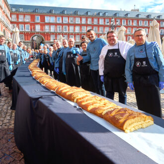 Fotografía para ilustrar la agenda cultural donde aparecen panaderos y pasteleros posan junto a una corona de la Almudena dispuesto sobre una larga mesa en la Plaza Mayor de Madrid, durante un evento festivo al aire libre.