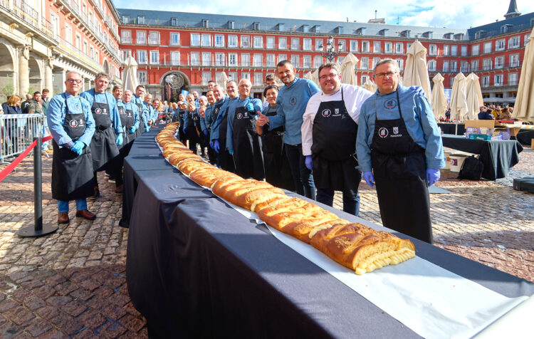 Fotografía para ilustrar la agenda cultural donde aparecen panaderos y pasteleros posan junto a una corona de la Almudena dispuesto sobre una larga mesa en la Plaza Mayor de Madrid, durante un evento festivo al aire libre.