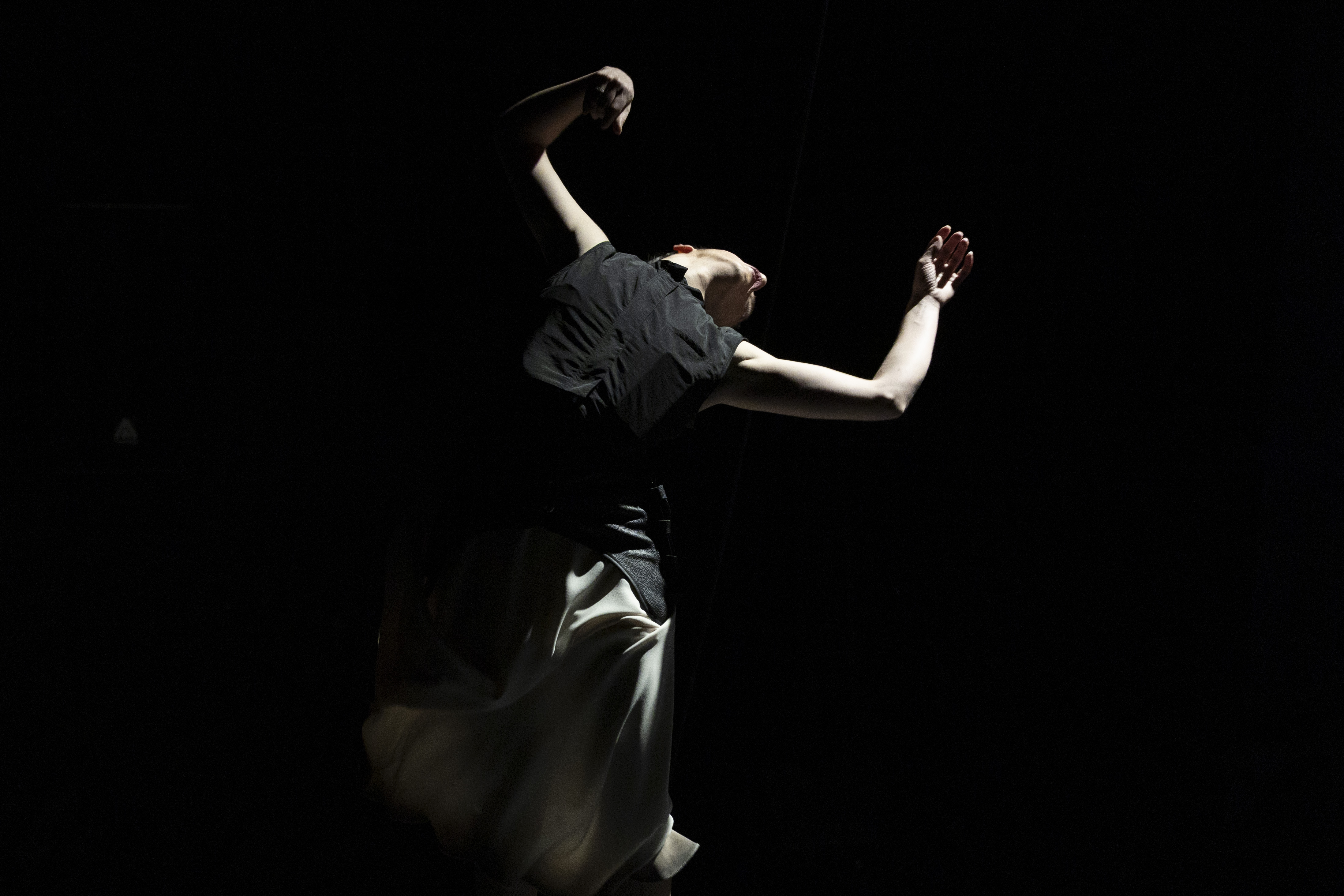 Fotografía artística de una bailarina en movimiento sobre un fondo negro, iluminada por un haz de luz que resalta sus brazos extendidos y su falda clara, creando un fuerte contraste entre luz y sombra.