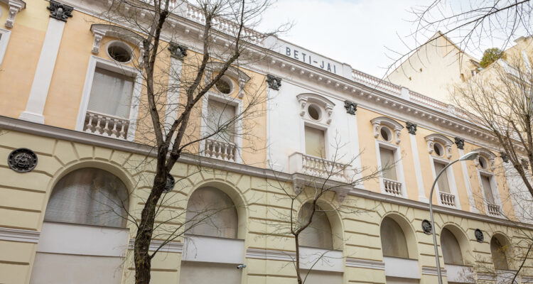 Fachada restaurada del frontón Beti Jai en Madrid, con elementos decorativos neoclásicos y árboles en primer plano.