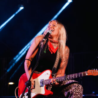 Foto promocional de Blanca Jurado donde se ve a la artista tocando la guitarra eléctrica y cantando, vestida con pantalón de leopardo, camiseta negra y pañuelo rojo al cuello