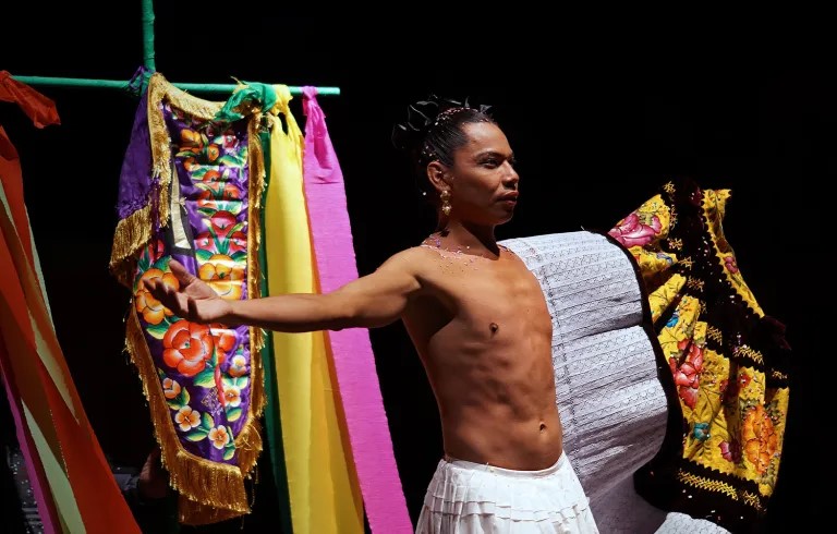 Fotografía escénica de un bailarín con el torso desnudo y los brazos extendidos, rodeado de telas coloridas y bordadas, en un ambiente teatral con fondo oscuro.