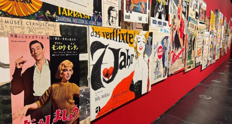 Muestra de la exposición sobre Marilyn Monroe donde se observa una pared roja cubierta por una larga hilera de carteles de cine antiguos en distintos idiomas.