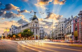 Imagen de la Gran Vía de Madrid al atardecer, donde se aprecia el edificio Metrópolis con su cúpula iluminada y un cielo parcialmente nublado con tonos dorados. En primer plano, las luces del tráfico en movimiento crean líneas de luz que refuerzan la sensación de dinamismo. La escena muestra uno de los puntos más representativos de la ciudad, un reflejo de la vitalidad, la actividad urbana y el atractivo que convierten a Madrid en un destino líder para el turismo de reuniones y congresos.