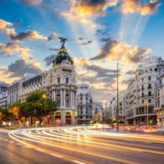 Imagen de la Gran Vía de Madrid al atardecer, donde se aprecia el edificio Metrópolis con su cúpula iluminada y un cielo parcialmente nublado con tonos dorados. En primer plano, las luces del tráfico en movimiento crean líneas de luz que refuerzan la sensación de dinamismo. La escena muestra uno de los puntos más representativos de la ciudad, un reflejo de la vitalidad, la actividad urbana y el atractivo que convierten a Madrid en un destino líder para el turismo de reuniones y congresos.