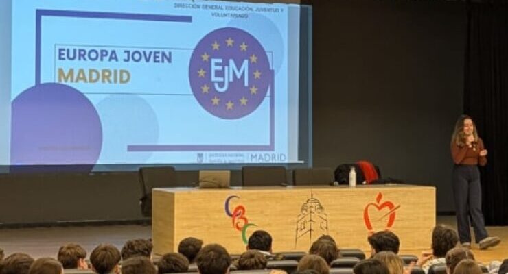 Ponente presentando Europa Joven Madrid en un auditorio escolar ante una audiencia de estudiantes, con proyección en pantalla grande y telón rojo de fondo