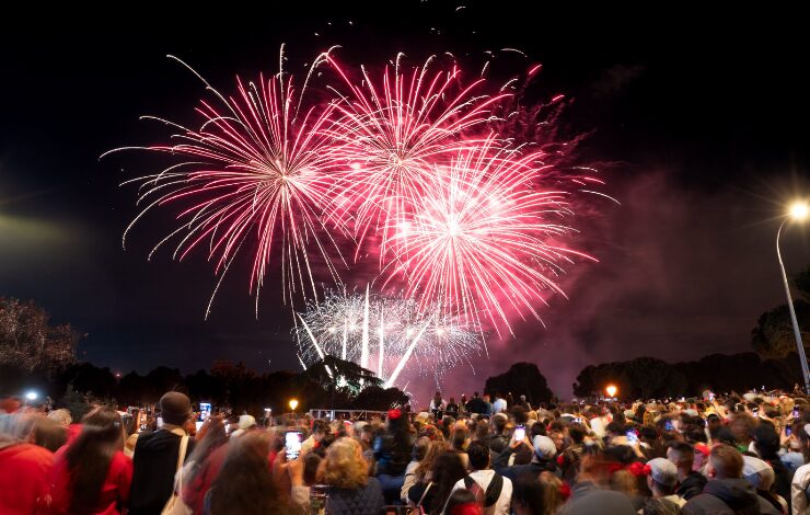 Fuegos artificiales iluminan el cielo nocturno mientras una multitud de personas observa y fotografía el espectáculo al aire libre durante una celebración festiva en Madrid