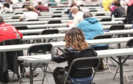 Esta imagen muestra a una multitud de personas participando en un examen masivo de oposiciones dentro de un gran salón de convenciones. Los asistentes están sentados individualmente en largas mesas blancas, manteniendo una organización rigurosa y uniforme durante la evaluación.