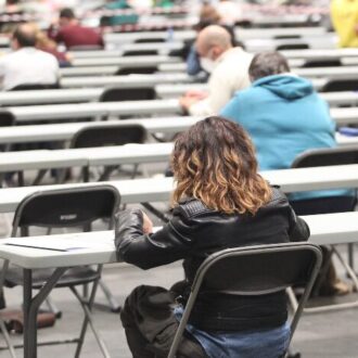 Esta imagen muestra a una multitud de personas participando en un examen masivo de oposiciones dentro de un gran salón de convenciones. Los asistentes están sentados individualmente en largas mesas blancas, manteniendo una organización rigurosa y uniforme durante la evaluación.