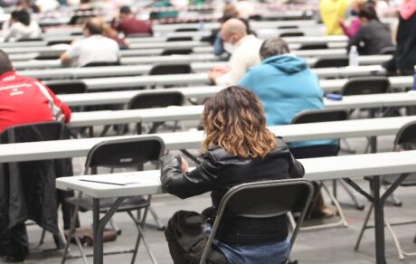 Esta imagen muestra a una multitud de personas participando en un examen masivo de oposiciones dentro de un gran salón de convenciones. Los asistentes están sentados individualmente en largas mesas blancas, manteniendo una organización rigurosa y uniforme durante la evaluación.