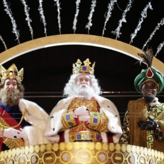 Melchor, Gaspar y Baltasar saludan desde una carroza dorada durante la Cabalgata de Reyes, vestidos con trajes reales y coronas, bajo un arco iluminado y fuegos fríos.