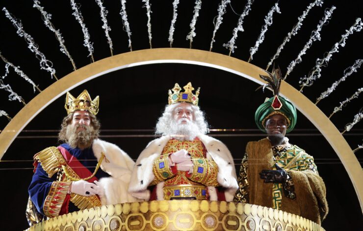 Melchor, Gaspar y Baltasar saludan desde una carroza dorada durante la Cabalgata de Reyes, vestidos con trajes reales y coronas, bajo un arco iluminado y fuegos fríos.