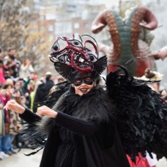 Persona disfrazada en un desfile de carnaval, vistiendo un traje negro con grandes alas de plumas y una máscara elaborada con detalles rojos y negros; detrás se ve una figura gigante con forma de criatura fantástica y una multitud observando en la calle.