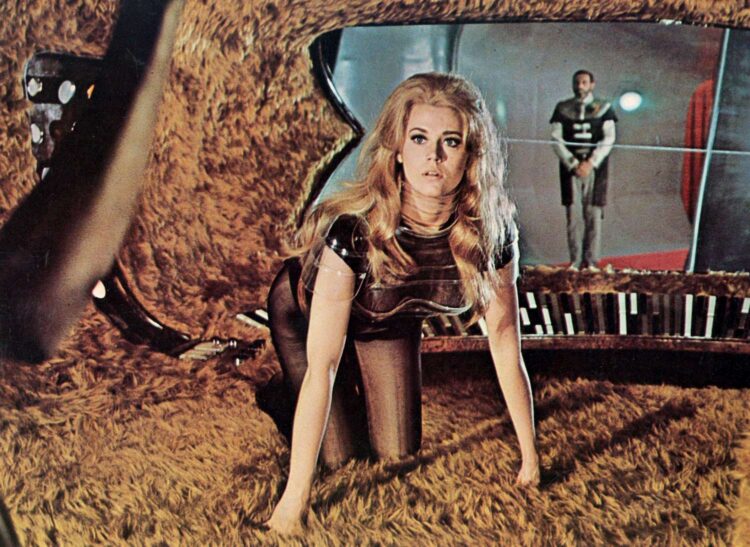Jane Fonda con traje futurista oscuro en un escenario de ciencia ficción cubierto de superficies peludas, con otra figura al fondo tras una mampara transparente.