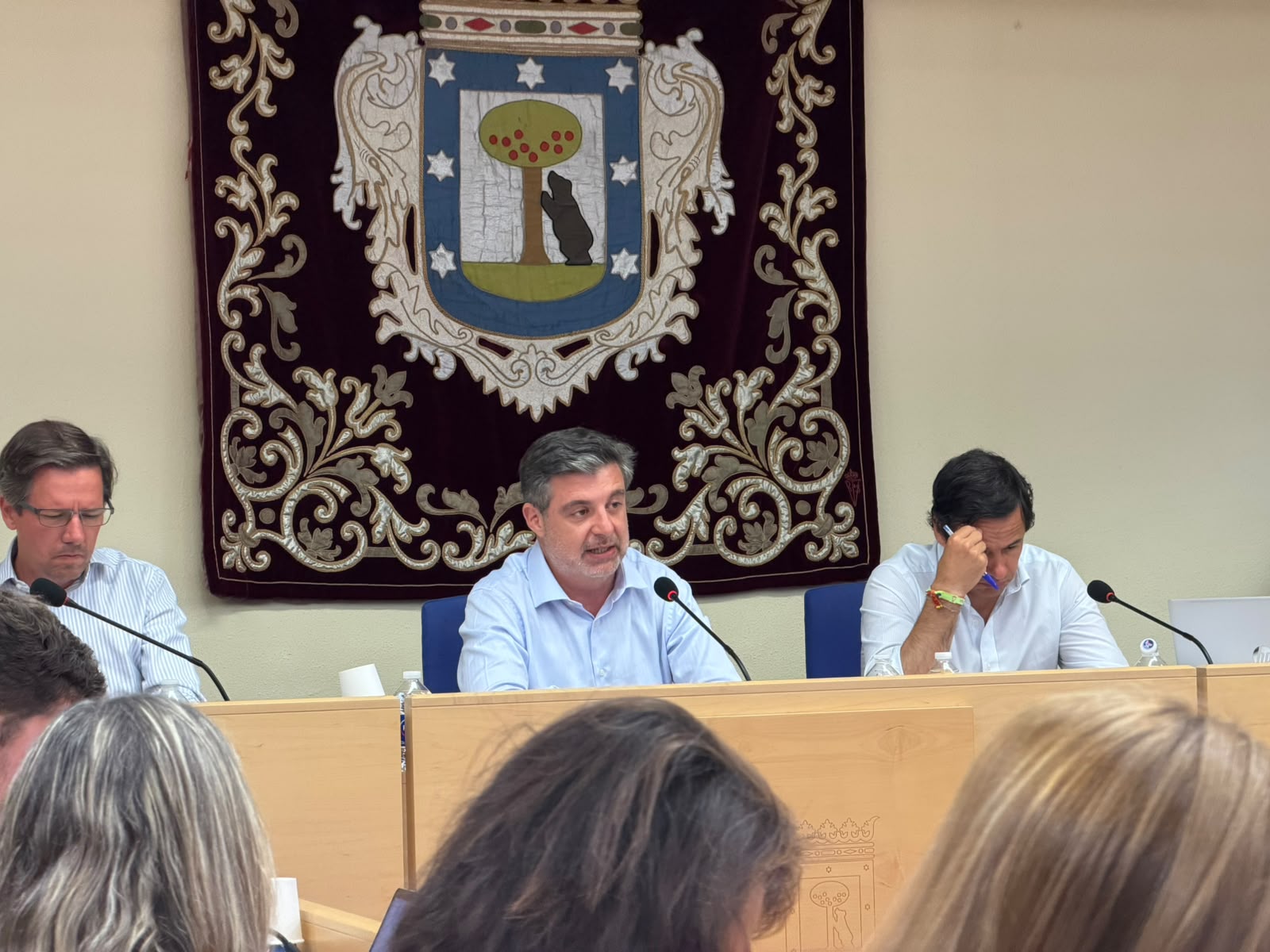 Intervención durante el debate del estado del distrito de Barajas en la Junta Municipal, con responsables municipales sentados en la mesa bajo el escudo oficial y público asistente en primer plano.