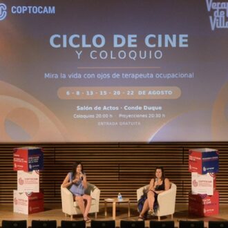 Dos ponentes sentadas en un escenario durante un coloquio, con una intérprete de lengua de signos a la derecha traduciendo la sesión. Al fondo, una pantalla anuncia el "Ciclo de Cine y Coloquio" de Coptocam dentro de Veranos de la Villa.