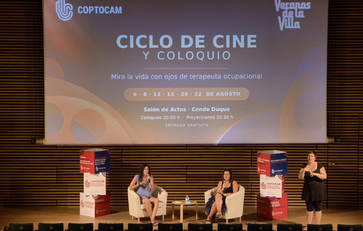 Dos ponentes sentadas en un escenario durante un coloquio, con una intérprete de lengua de signos a la derecha traduciendo la sesión. Al fondo, una pantalla anuncia el "Ciclo de Cine y Coloquio" de Coptocam dentro de Veranos de la Villa.