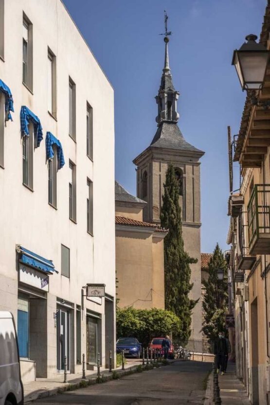 Calle del casco histórico de Fuencarral con la torre campanario de la iglesia al fondo, entre edificios de fachadas claras y un ciprés